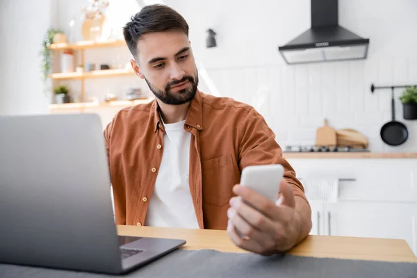 Bearded freelancer holding smartphone near laptop while working from home — Stock Photo