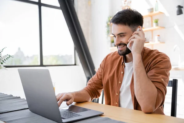 Alegre freelancer hablando en el teléfono inteligente cerca del ordenador portátil mientras trabaja desde casa - foto de stock