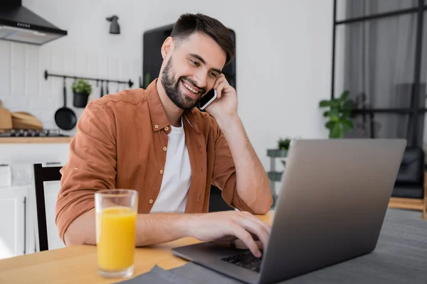 Joyful freelancer talking on smartphone near laptop while working from home — Stock Photo