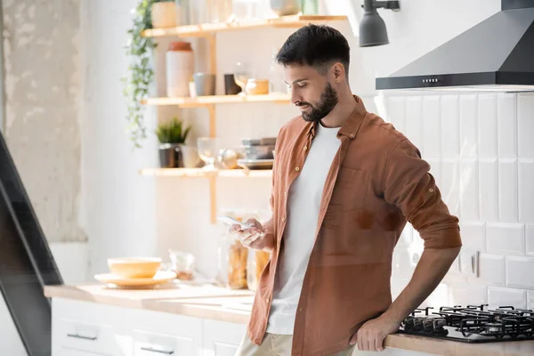 Mensajería hombre barbudo en el teléfono inteligente mientras está de pie cerca de la estufa en la cocina - foto de stock