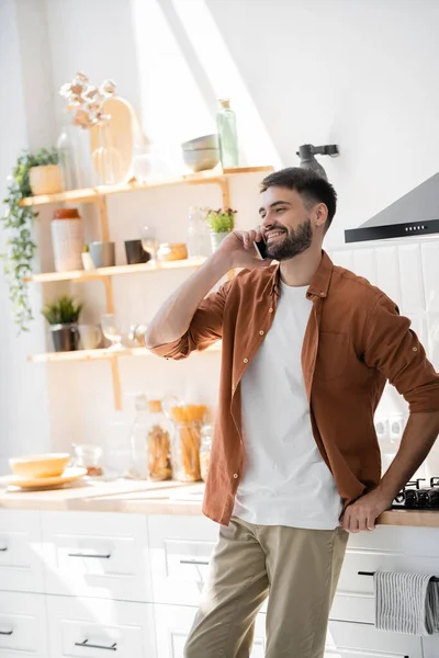 Happy bearded man talking on smartphone while standing in modern kitchen — Stock Photo