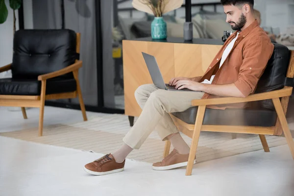 Полная длина бородатый фрилансер сидит на кожаном кресле и с помощью ноутбука — стоковое фото