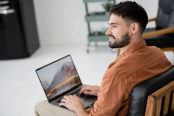 Cheerful and bearded freelancer sitting on leather armchair with modern laptop — Stock Photo