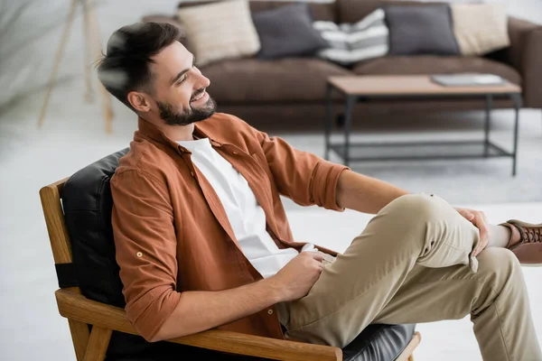 Hombre alegre y barbudo sentado en un cómodo sillón de cuero en la sala de estar - foto de stock