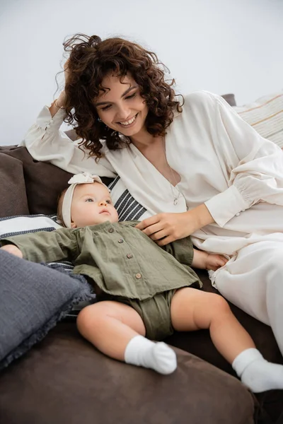 Felice madre in abito sorridente vicino figlia neonata in fascia seduta sul divano — Foto stock