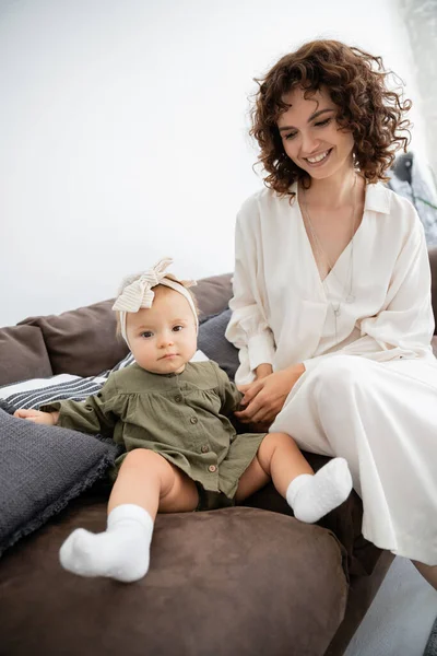 Веселая мать в платье улыбается рядом с младенческой дочерью в повязке на голове сидя на диване — стоковое фото