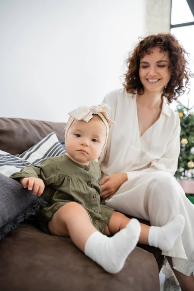 Feliz madre en vestido sonriendo mientras mira a la niña sentada en el sofá - foto de stock