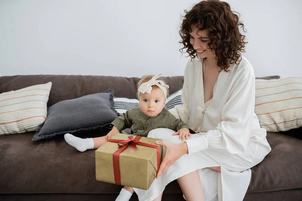 Happy mother in dress holding wrapped gift box near infant daughter in headband sitting on sofa — Stock Photo