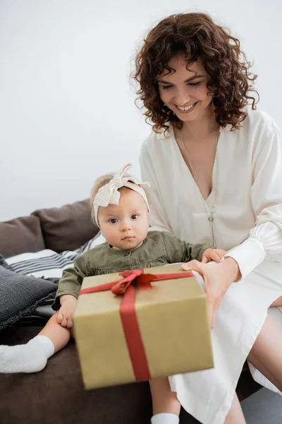 Весела мати в сукні тримає загорнуту подарункову коробку біля немовляти дочка в пов'язці, сидячи на дивані — стокове фото