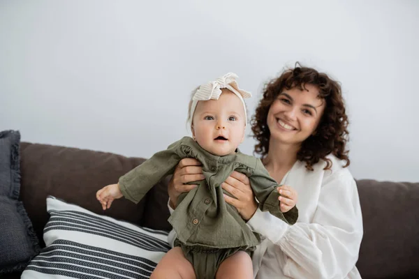 Mère heureuse avec les cheveux bouclés souriant tout en tenant dans les bras fille bébé dans le bandeau — Photo de stock