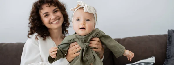 Felice madre con i capelli ricci sorridenti mentre tiene in braccio la figlia neonata in fascia, striscione — Foto stock