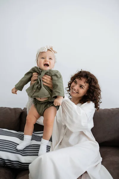 Щаслива мати з кучерявим волоссям посміхається, тримаючись в руках збуджена дочка-немовля в пов'язці — стокове фото