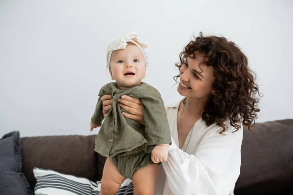 Feliz madre con el pelo rizado sonriendo mientras sostiene en brazos bebé hija en vestido y diadema - foto de stock