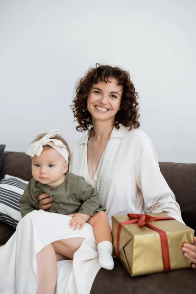 Довольная и кудрявая мать в платье с обернутой подарочной коробкой рядом с младенческой дочерью в повязке — стоковое фото