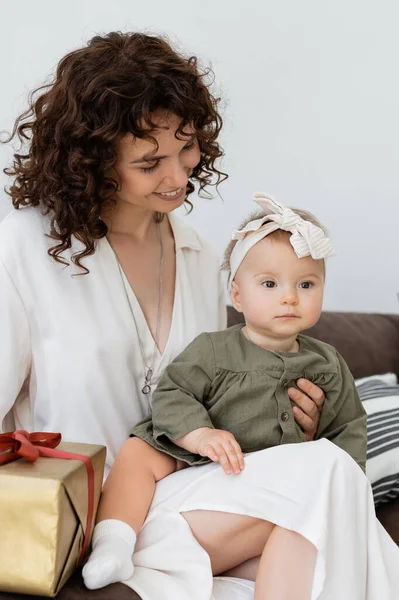 Счастливая и кудрявая мать в платье с завернутой подарочной коробкой рядом с младенческой дочерью в повязке — стоковое фото