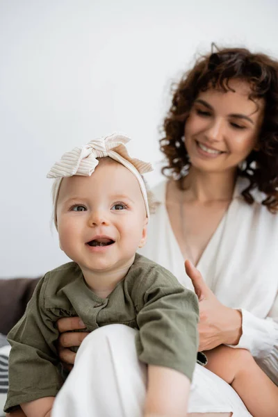 Glückliche Mutter mit lockigem Haar lächelt, während sie das kleine Mädchen im Stirnband im Arm hält — Stockfoto