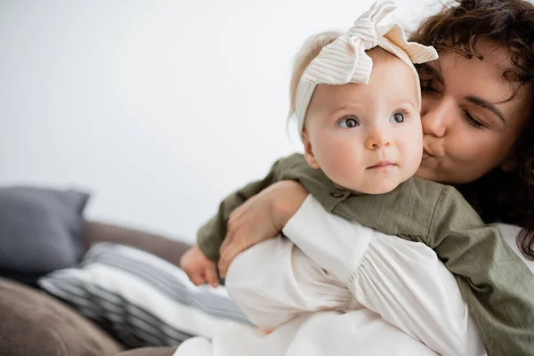 Curly mother hugging and kissing cheek of baby girl in headband — Stock Photo