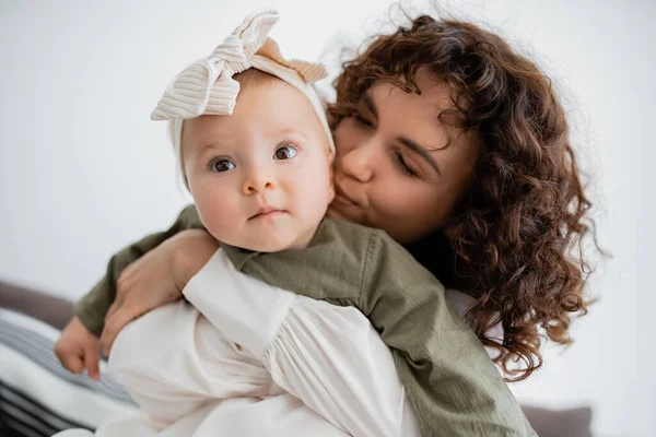 Кудрявая мать обнимает и целует щеку младенца в повязке на голове — стоковое фото