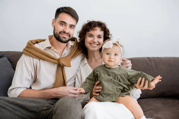 Fröhliche Eltern und kleines Mädchen blicken in die Kamera, während sie auf dem Sofa im Wohnzimmer sitzen — Stockfoto