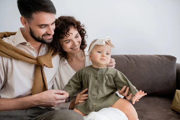 Genitori allegri guardando la bambina in fascia mentre si siede sul divano in soggiorno — Foto stock