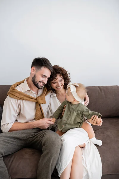 Positive parents looking at infant girl in headband while sitting on sofa in living room — Stock Photo