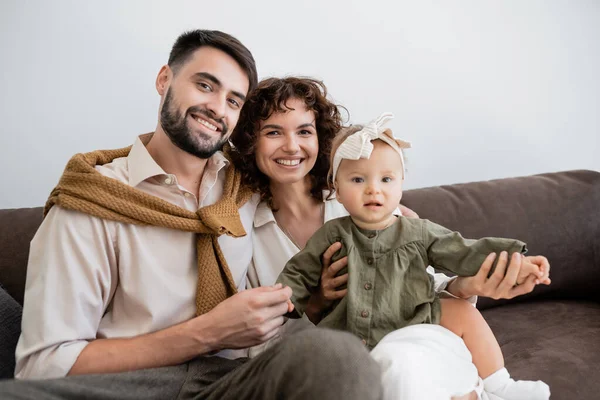 Padres positivos y niña mirando a la cámara mientras se sienta en el sofá en la sala de estar - foto de stock