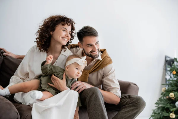 Padres positivos sosteniendo bebé niña en diadema y sonriendo cerca borrosa árbol de Navidad - foto de stock