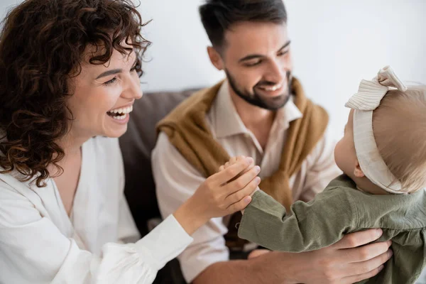 Animado marido e mulher segurando bebê menina na cabeça e sorrindo na sala de estar — Fotografia de Stock