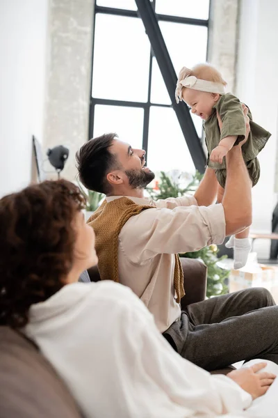 Cheerful father holding infant daughter near blurred wife in living room — Stock Photo
