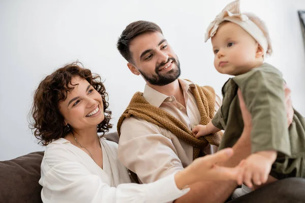 Mari et femme joyeux tenant bébé fille dans le bandeau et souriant dans le salon — Photo de stock