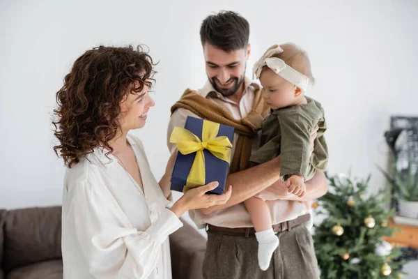 Mãe feliz segurando caixa de presente perto do marido alegre segurando a filha infantil no Natal — Fotografia de Stock