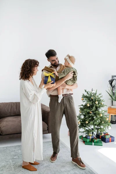 Feliz madre sosteniendo caja de regalo cerca de marido sonriente sosteniendo hija bebé en Navidad - foto de stock