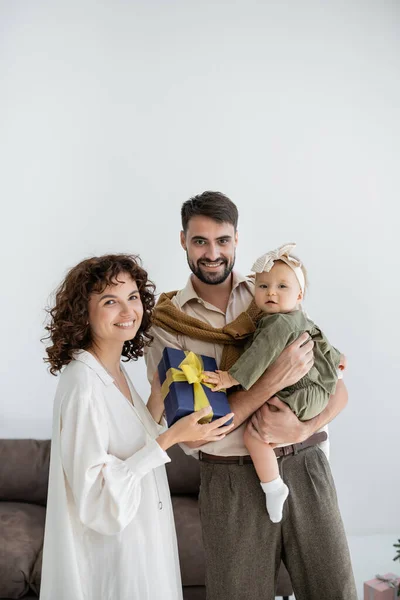 Glückliche Mutter hält Geschenkbox neben lächelndem Ehemann, der kleine Tochter in den Armen hält — Stockfoto