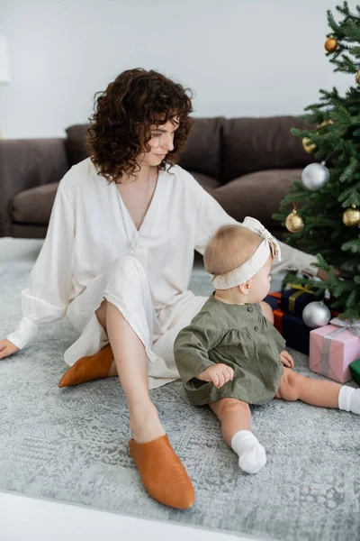 Lockige Mutter sitzt neben geschmücktem Weihnachtsbaum mit Geschenken und kleiner Tochter — Stockfoto