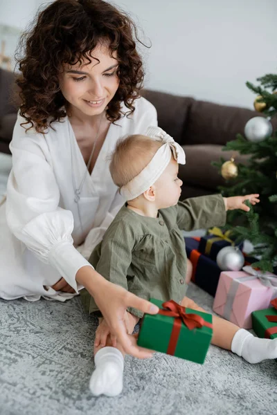 Alegre madre sosteniendo caja de regalo cerca de árbol de Navidad decorado e hija bebé - foto de stock