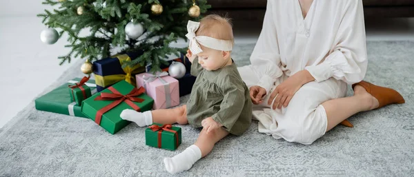 Mutter sitzt neben Säugling Tochter schaut auf Geschenkbox neben geschmücktem Weihnachtsbaum, Banner — Stockfoto