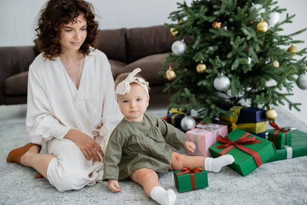 Joyeuse mère assise près de bébé fille et présente sous l'arbre de Noël décoré — Photo de stock