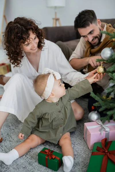 Menina infantil curioso tocando bola de Natal no pinho decorado perto de pais felizes — Fotografia de Stock