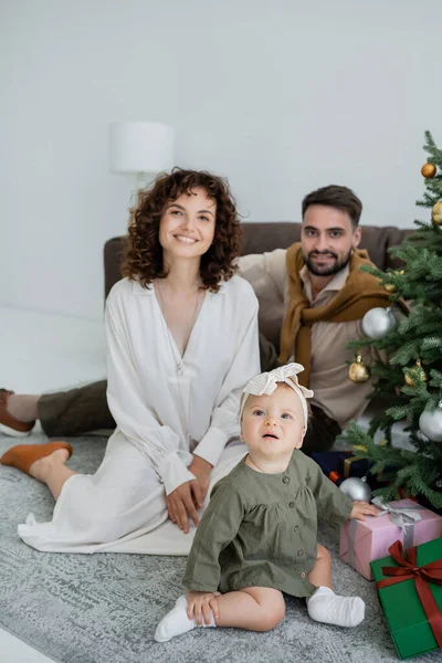 Cheerful family with infant baby girl sitting near christmas tree with presents — Stock Photo