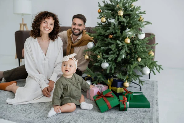 Joyeuse famille avec bébé fille assise près de l'arbre de Noël avec des cadeaux — Photo de stock