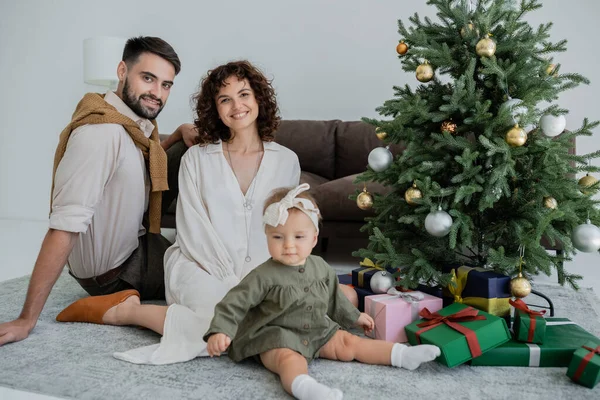 Padres felices con niña sentada cerca del árbol de Navidad con regalos - foto de stock