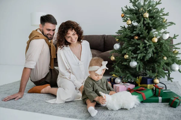 Genitori felici guardando neonata seduta vicino all'albero di Natale con regali — Foto stock