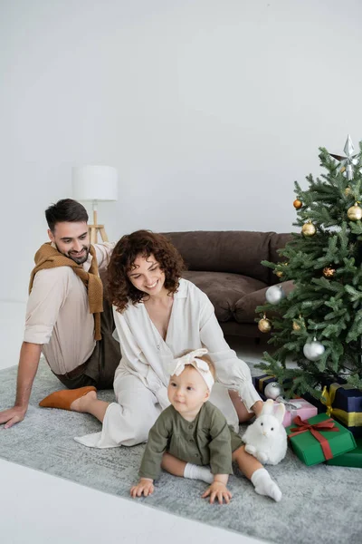 Genitori felici e neonata seduta vicino all'albero di Natale decorato con regali — Foto stock