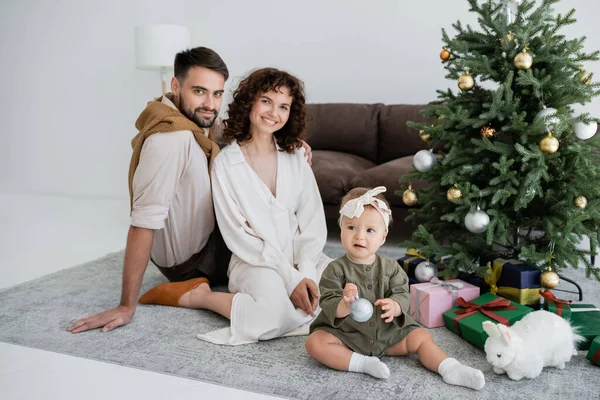 Padres felices y niña sentada cerca del árbol de Navidad decorado con regalos - foto de stock