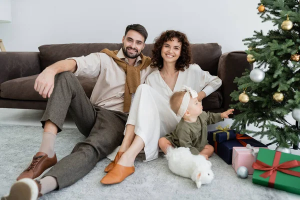 Happy parents sitting near infant girl and decorated christmas tree with presents — Stock Photo