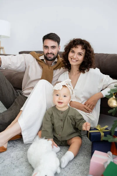 Padres felices y niña alegre sentado cerca del árbol de Navidad decorado con regalos - foto de stock