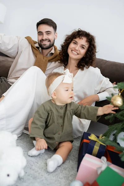 Любопытная малышка трогает рождественский бал на украшенной сосне рядом с веселыми родителями — стоковое фото