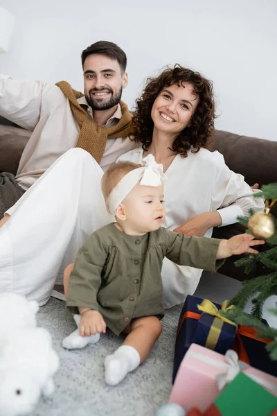 Curious infant girl touching christmas ball on decorated pine near cheerful parents — Stock Photo