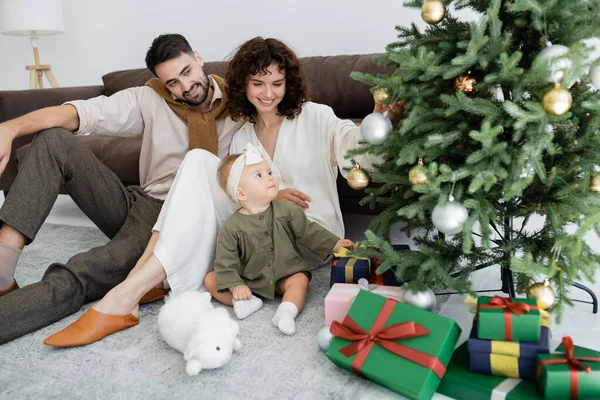 Pais felizes e menina infantil sentado perto de caixas de presente sob árvore de natal decorada — Fotografia de Stock