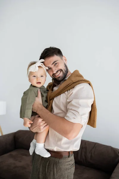 Homme barbu positif tenant dans les bras bébé fille en robe et bandeau — Photo de stock